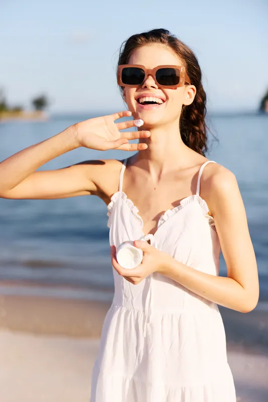 Lively beach scene featuring a young woman in a white sundress and sunglasses, smiling as she holds a round object near her chest, radiating carefree summer joy.