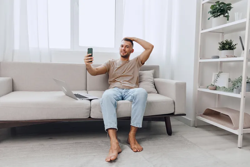 Smiling man taking selfie in cozy living room while sitting on sofa, representing digital detox and soft masculinity in a relaxed atmosphere.
