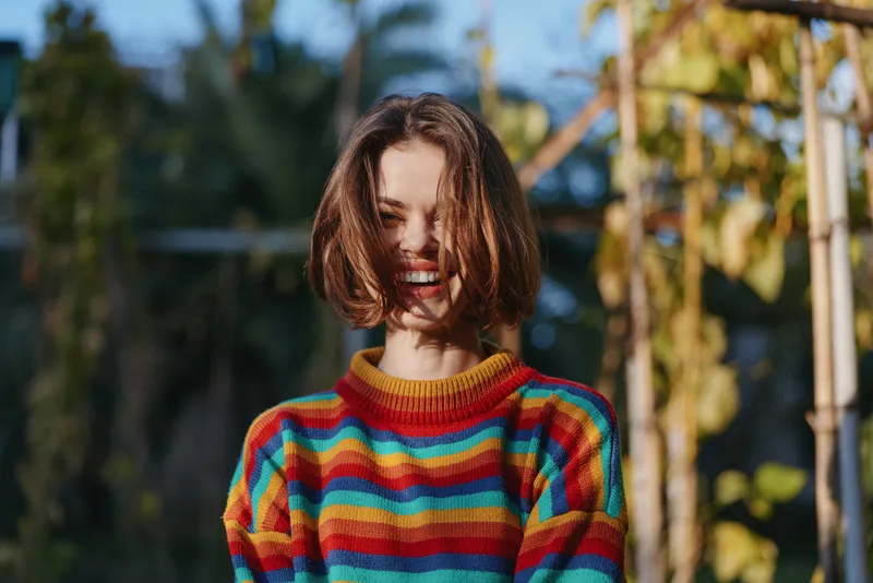 Woman smile portrait sweater short hair outdoors, happy young woman laughing in colorful striped knit sweater, candid natural light, joyful casual fashion and lifestyle moment.