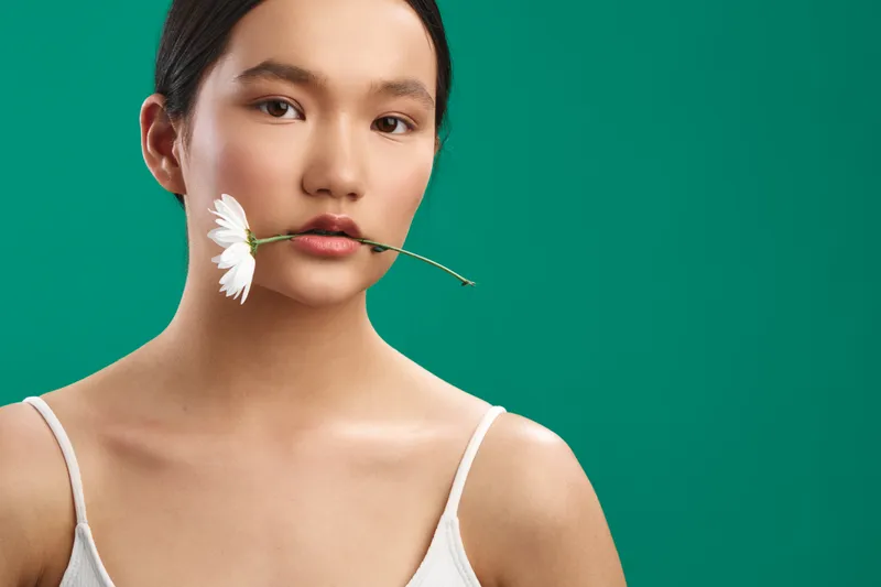 asian woman holds a white flower between her lips against a vivid green background. minimalist portrait with soft lighting and clean composition, conveying calm elegance and natural beauty.