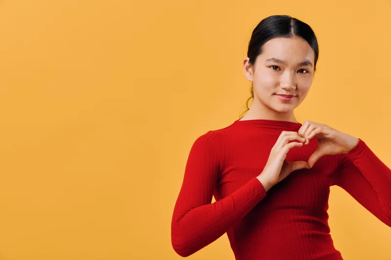 Young woman in red sweater making heart shape with hands, smiling and posing against yellow background. Female model, gesture of love or affection, studio portrait with positive emotion.
