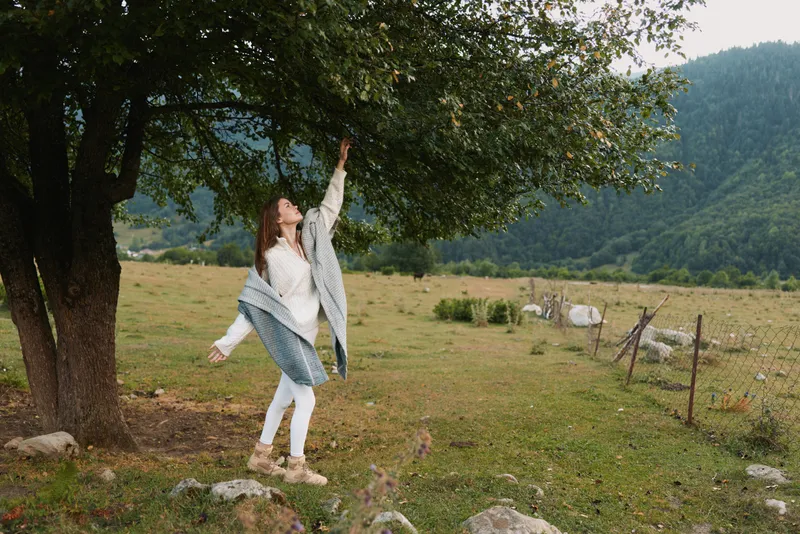 A woman reaches up to a branch in a pastoral field, blending with a rustic landscape and grazing cows in the background for a moment of nature connection.