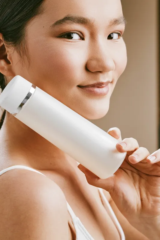 asian woman holds a white bottle close to her face, conveying skincare focus with a gentle smile and calm mood in a clean, minimal setting.
