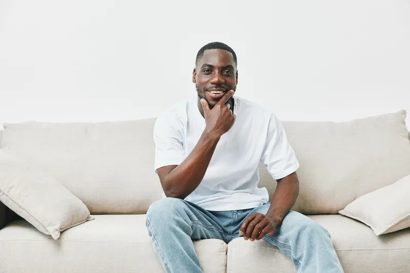 Young Black man in casual wear smiling thoughtfully on a light colored couch, conveying confidence and relaxation in a cozy living space