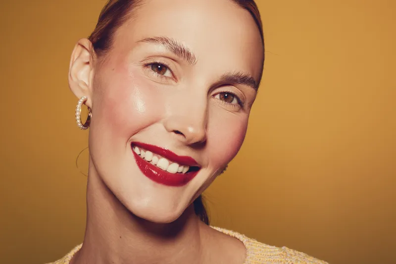 Close-up portrait of smiling woman with red lipstick and hoop earrings on yellow background. Concept of beauty, makeup, happiness, and confident female model.