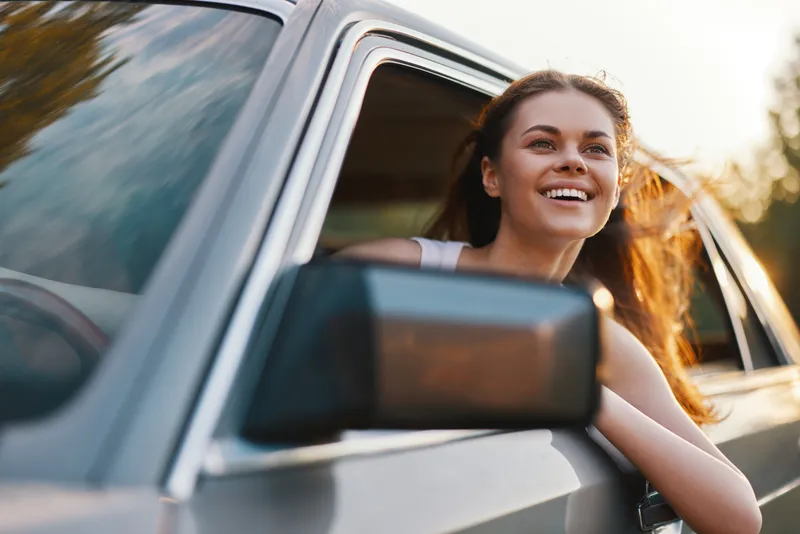 Young woman with long hair enjoying a road trip, smiling joyfully while leaning out of a car window during sunset, embodying freedom and adventure.