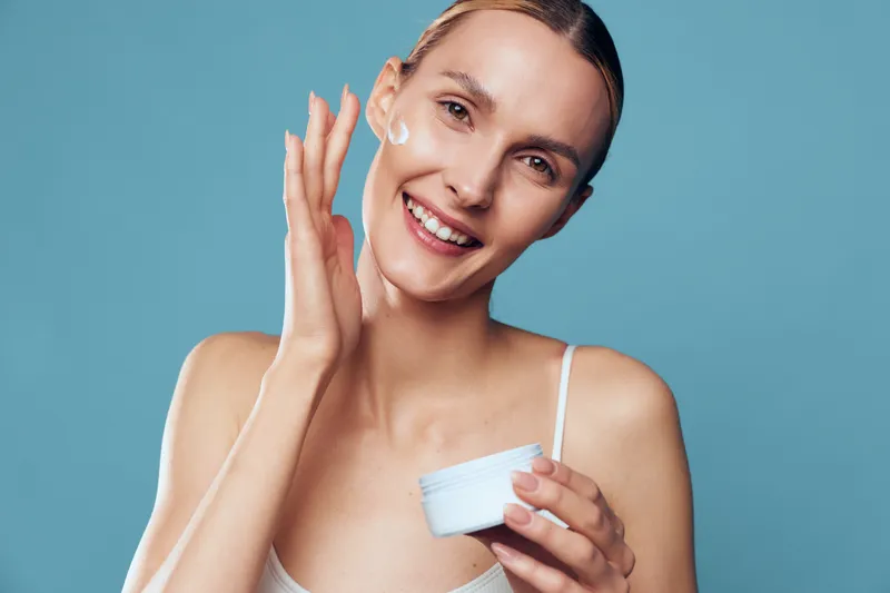 Smiling woman applies face cream on cheek holding white jar skincare moisturizer with clean fresh skin isolated on blue background beauty care concept natural look.