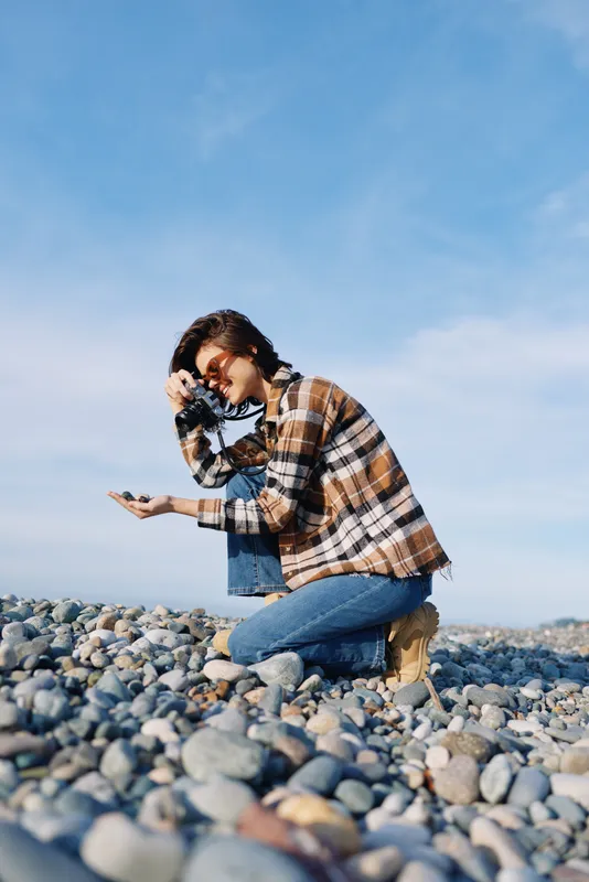 Woman kneeling on a pebbled beach in a plaid shirt, exploring stones under a clear blue sky in an outdoor scene, casual attire and curious pose captured in natural light.