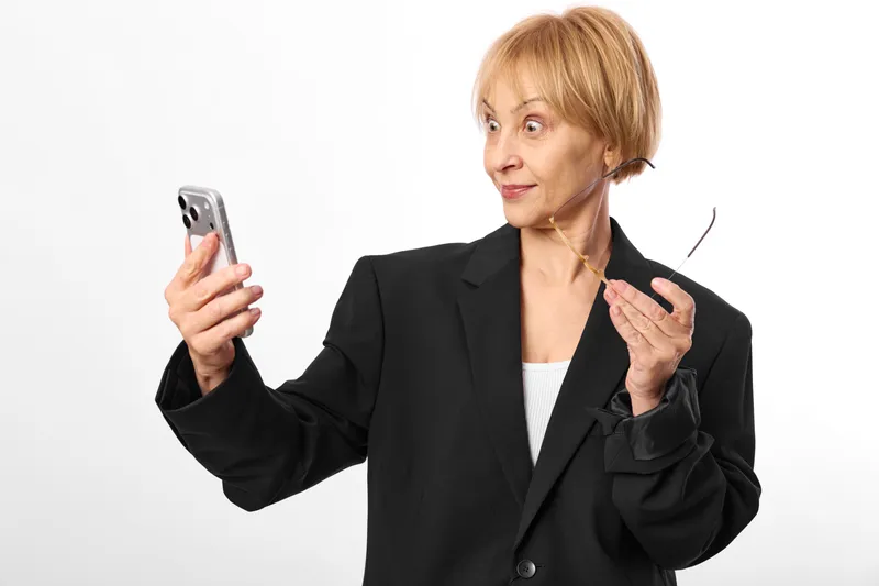 Woman holding smartphone and earbuds, surprised expression, wearing business suit. Indoors studio portrait about communication technology and headset listening mood, modern concept.