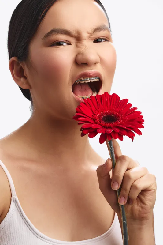 Asian woman poses closely with a vibrant red flower held near her open mouth, conveying playful boldness in a bright studio shot with soft, flattering lighting.