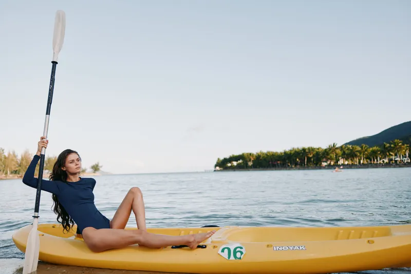 Woman in a blue swimsuit lounges on a yellow kayak on calm water, paddle upright, sunny sky, serene leisure moment on a scenic lake.