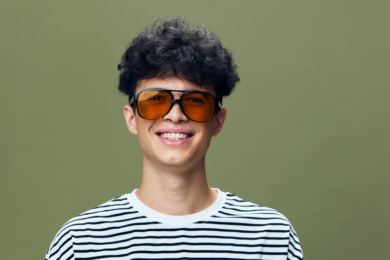 smiling man with curly hair wearing orange sunglasses and a striped shirt, posing against an olive green background, casual and youthful style