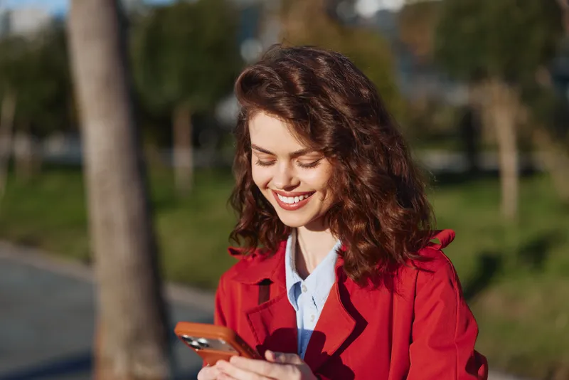 A young woman in a red coat smiles while looking at her phone outdoors in a sunny park, capturing a casual, everyday moment.