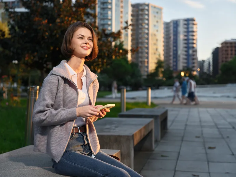 A cheerful woman sits on a concrete bench in a modern urban park, smiling while texting on a smartphone, with tall buildings in the background and warm daylight.