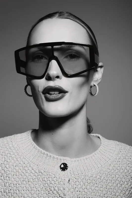 Portrait of a white woman with pale skin wearing large fashion sunglasses and hoop earrings. Studio lighting highlights modern style and confident expression.
