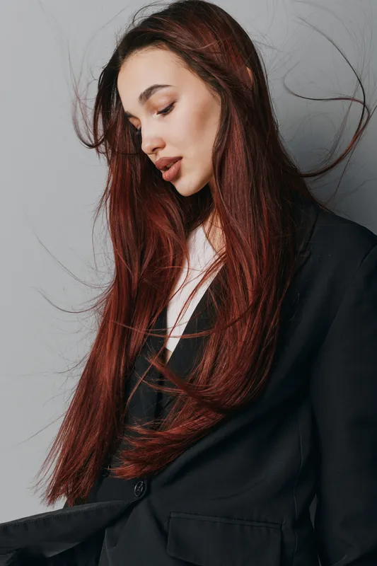 Woman with long red hair wearing black blazer posing with closed eyes and serene expression against gray background in studio. Beauty, fashion, portrait and style concept.