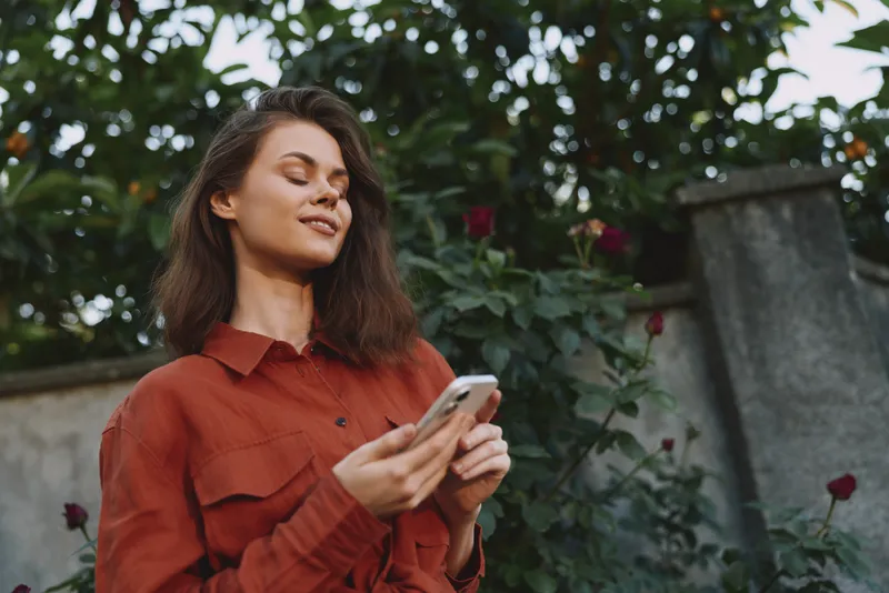 Young woman in a rust shirt outdoors, smiling with her eyes closed while checking her smartphone, enjoying a calm moment in a lush green setting with natural colors