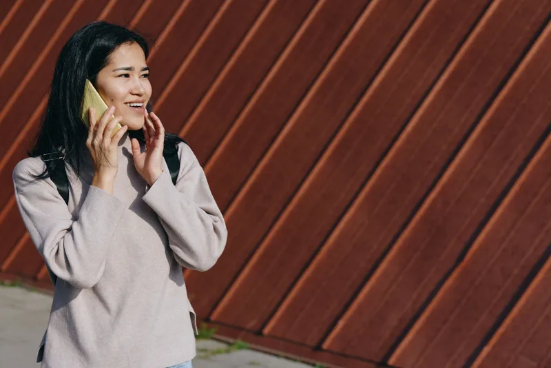 Woman talking on a mobile phone outdoors beside a wooden wall, casual sweater, sunlit candid moment, urban lifestyle and everyday communication in a street setting