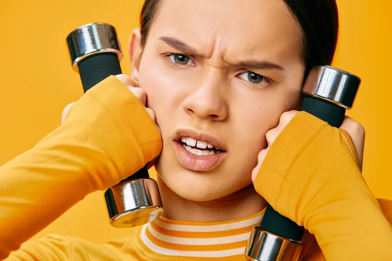 Fitness woman with dumbbells pressed up to her cheeks in a bold orange studio, intense expression during a workout, capturing focus, strength and energetic training.