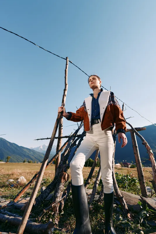 Person standing outdoors in field by wooden fence holding stick, wearing boots and brown shearling jacket with white pants, framed against mountains and clear blue sky.