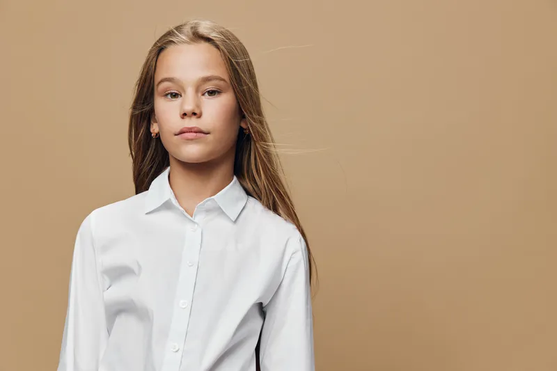 Girl portrait in white shirt with long hair standing on beige background looking confident and calm with natural light in studio for childhood and fashion concept.