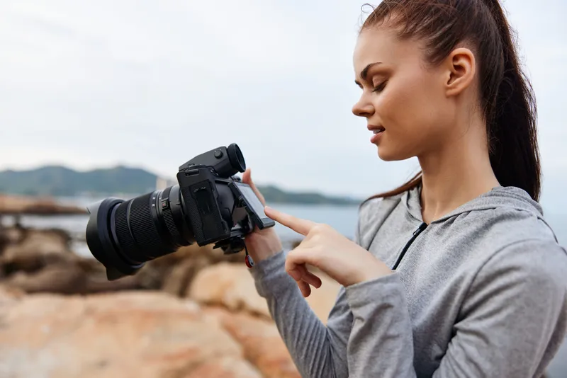 A young woman with long hair in a gray sports jacket checks her camera settings on a rocky beach, showcasing her passion for photography and nature
