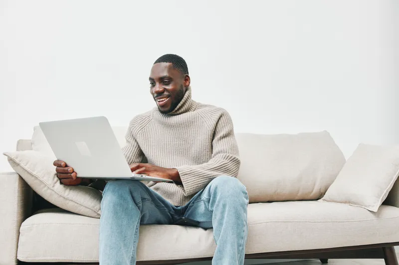 A joyful young Black man in a cozy sweater working on a laptop while sitting on a stylish light sofa, showcasing a modern and relaxed home environment