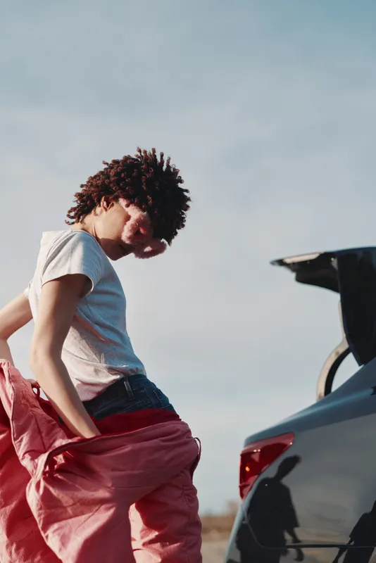 Casual man adjusting pink pants beside an open car trunk in a sunny outdoor setting, showcasing relaxed lifestyle and street style with attention to detail