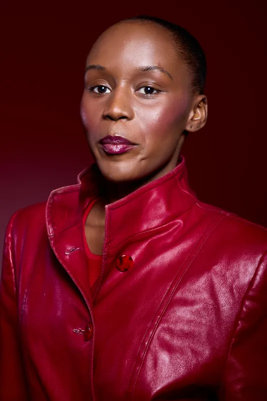 Confident African woman with dark skin wearing a red leather jacket poses against a deep red background. Studio portrait with elegant fashion and strong expression concept.