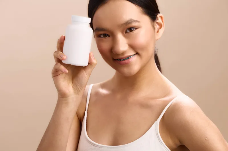 Asian woman smiles while holding a white bottle in a simple studio setting, presenting a skincare product with confidence and charm, suggesting daily care and wellness in a bright, approachable vibe.
