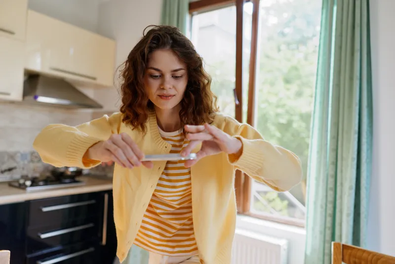 Young woman in a bright kitchen measures ingredients with a wooden spoon, wearing a yellow cardigan. Cozy home atmosphere, everyday cooking, warm natural light, candid lifestyle moment.