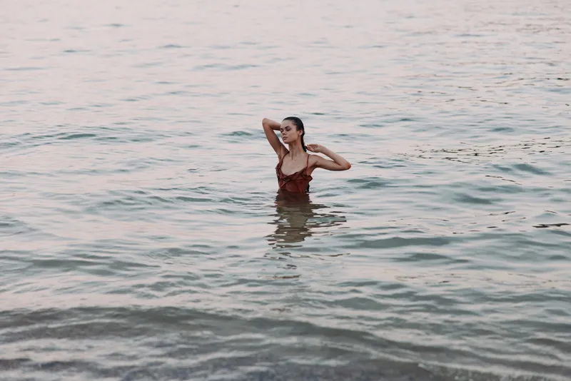 Young woman in red swimsuit standing in calm water, enjoying serene nature with relaxed and peaceful expression at sunset.