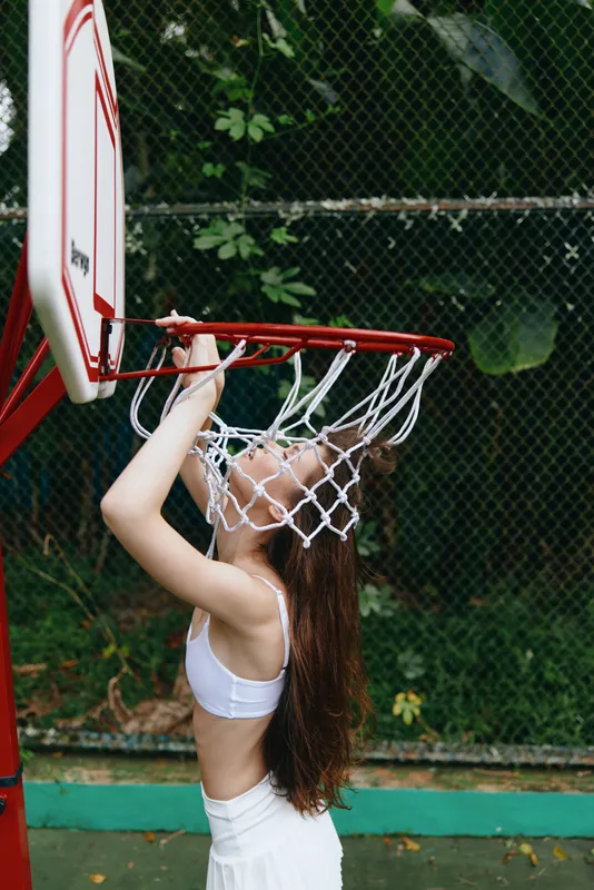 Young female athlete celebrating victory by hanging on a basketball hoop in a tropical outdoor setting, showcasing joy and confidence