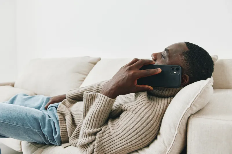 Young Black man relaxing on a sofa while talking on his smartphone, dressed in a cozy beige sweater, exuding a calm and content attitude in a minimalist living space