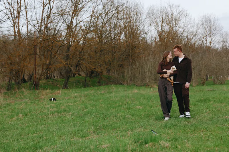 Smiling couple walking in a grassy park, casual outfits, gentle conversation, and a small pet cradled in arms. Natural daylight, relaxed lifestyle, and affectionate moment captured in an outdoor