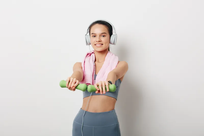 Fitness oriented woman in athletic wear, wearing headphones and towel around neck, holding green dumbbells for a home workout against a plain backdrop. Focus on health, strength, and motivation.