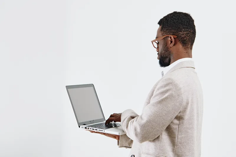 Professional young African American man in beige blazer and glasses working on laptop computer isolated on plain white background studio portrait. Business, technology, focus, professional lifestyle c