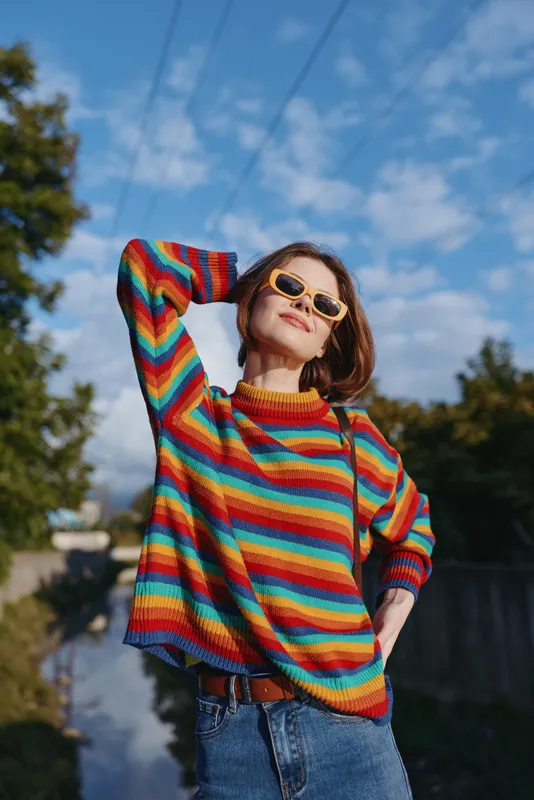 Woman in a colorful striped sweater and sunglasses outdoors, smiling in a casual outfit with denim jeans, short hair and relaxed pose in sunny blue sky lifestyle portrait for fashion travel.