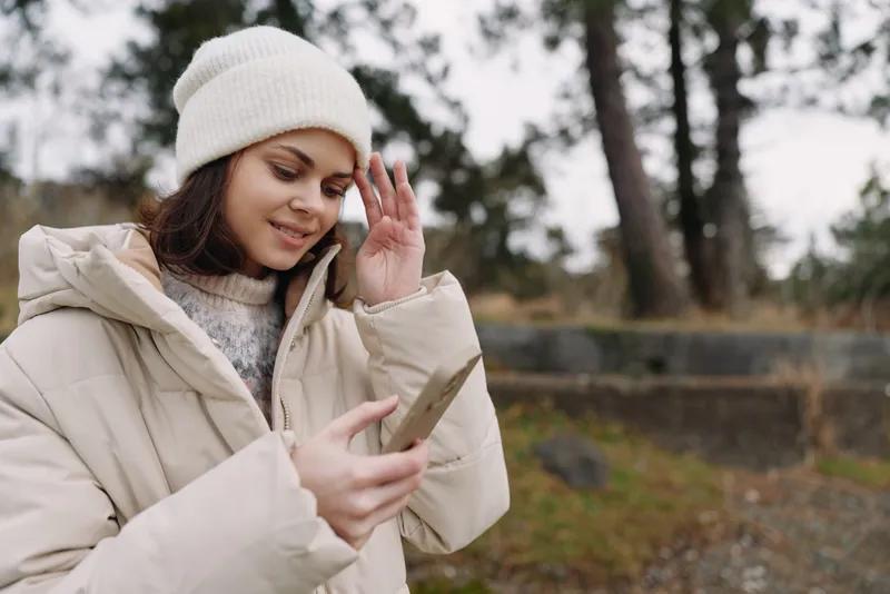 Woman in a warm winter coat and beanie using a phone outdoors, with a gentle smile and natural light in a quiet park setting.