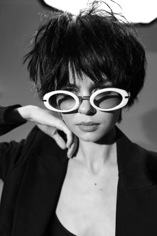 Black and white portrait of a stylish woman wearing round sunglasses and dark clothing with short hair.