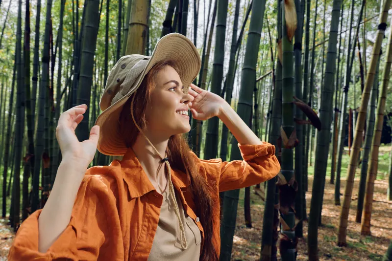 Woman wearing hat in a bamboo forest, portrait and smile as she enjoys nature and outdoor exploration; young female in casual jacket soaking sunlight in green grove.