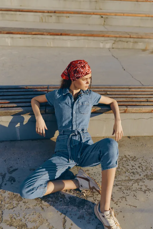 lifestyle woman wearing modern boho-western denim jumpsuit and red bandana sitting relaxed on concrete stairs in sunlit street style with film color tones