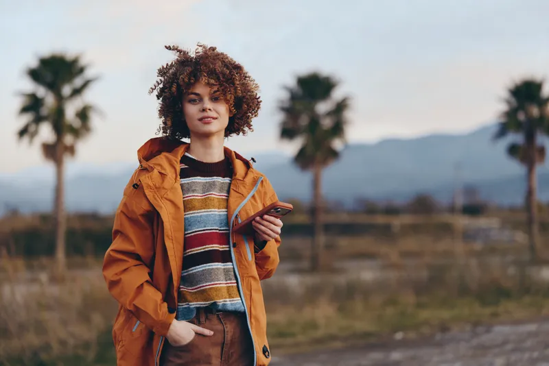 Woman lifestyle portrait outdoors, curly hair, orange jacket, striped sweater, standing on a rural road, palm trees and mountains in sunset light, relaxed travel mood