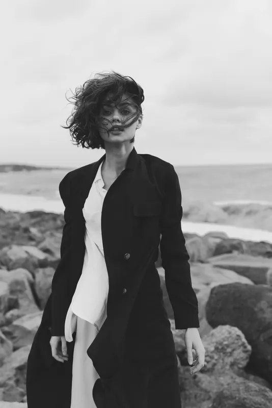 Windy coastal scene with a fashionable woman in a long dark coat walking along rocks, sea in the distance, moody monochrome tones, windblown hair and dramatic ocean atmosphere, emphasizing style and