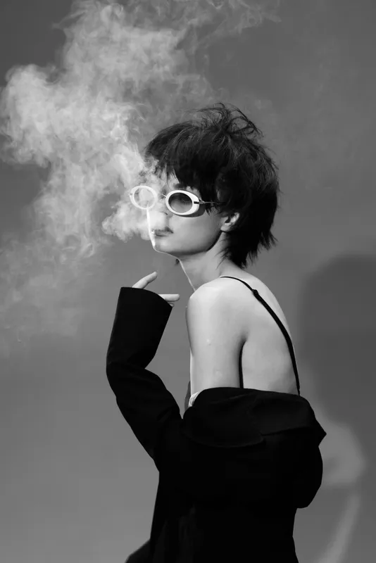 Black and white portrait of a stylish woman with sunglasses smoking against dark background.