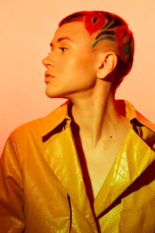 young woman with short colorful hair and floral shaved design wearing yellow textured jacket in warm orange lighting, creative hairstyle and modern fashion style