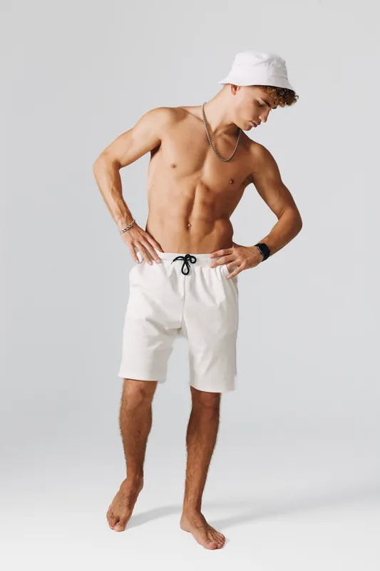 Young man with muscular body wearing white shorts and bucket hat posing barefoot on gray background with casual style and accessories for summer fashion.