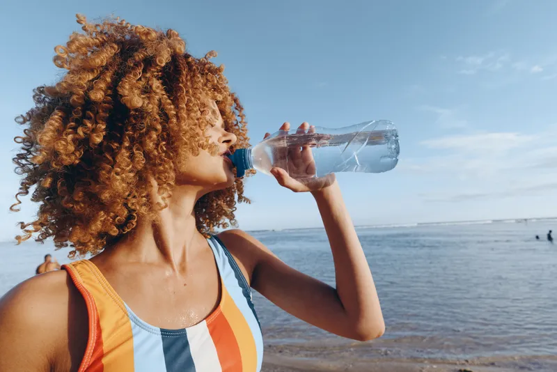 Woman curly hair drinking water bottle beach sea summer, close profile outdoors, clear container sip, bright sunny horizon light, wind and natural tones, wellness moment, calm expression