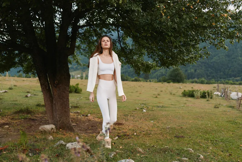 Young woman in white outfit walking outdoors near tree in grassy field with mountains in background natural lifestyle casual confident relaxed mood