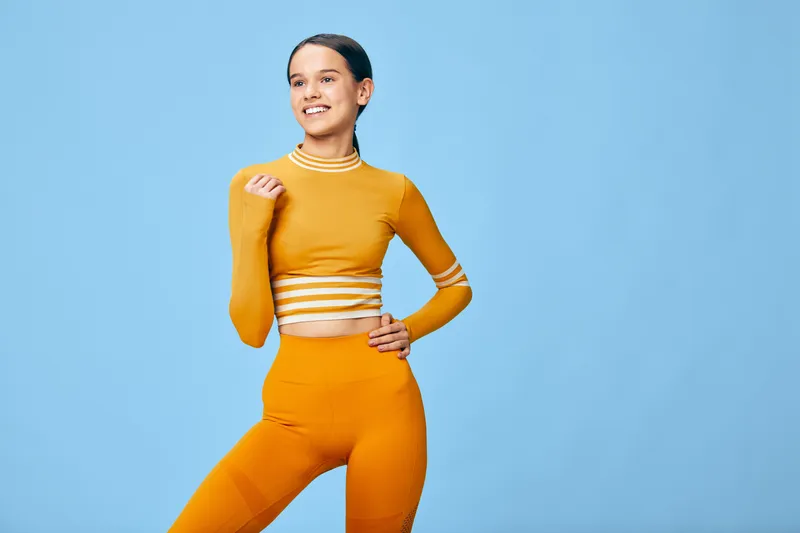 Woman in orange fitness outfit strikes a confident pose against a blue backdrop, showcasing athletic style, strength and balance during a workout session.